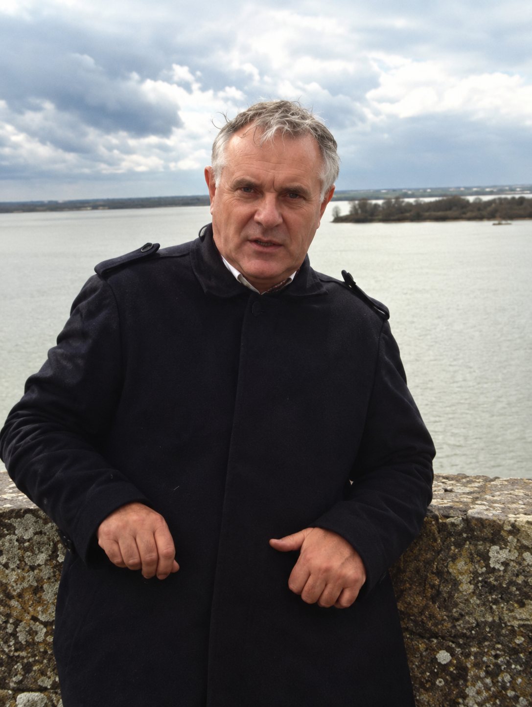 Un homme aux cheveux gris, vêtu d'un manteau sombre, pose devant un large fleuve sous un ciel couvert.