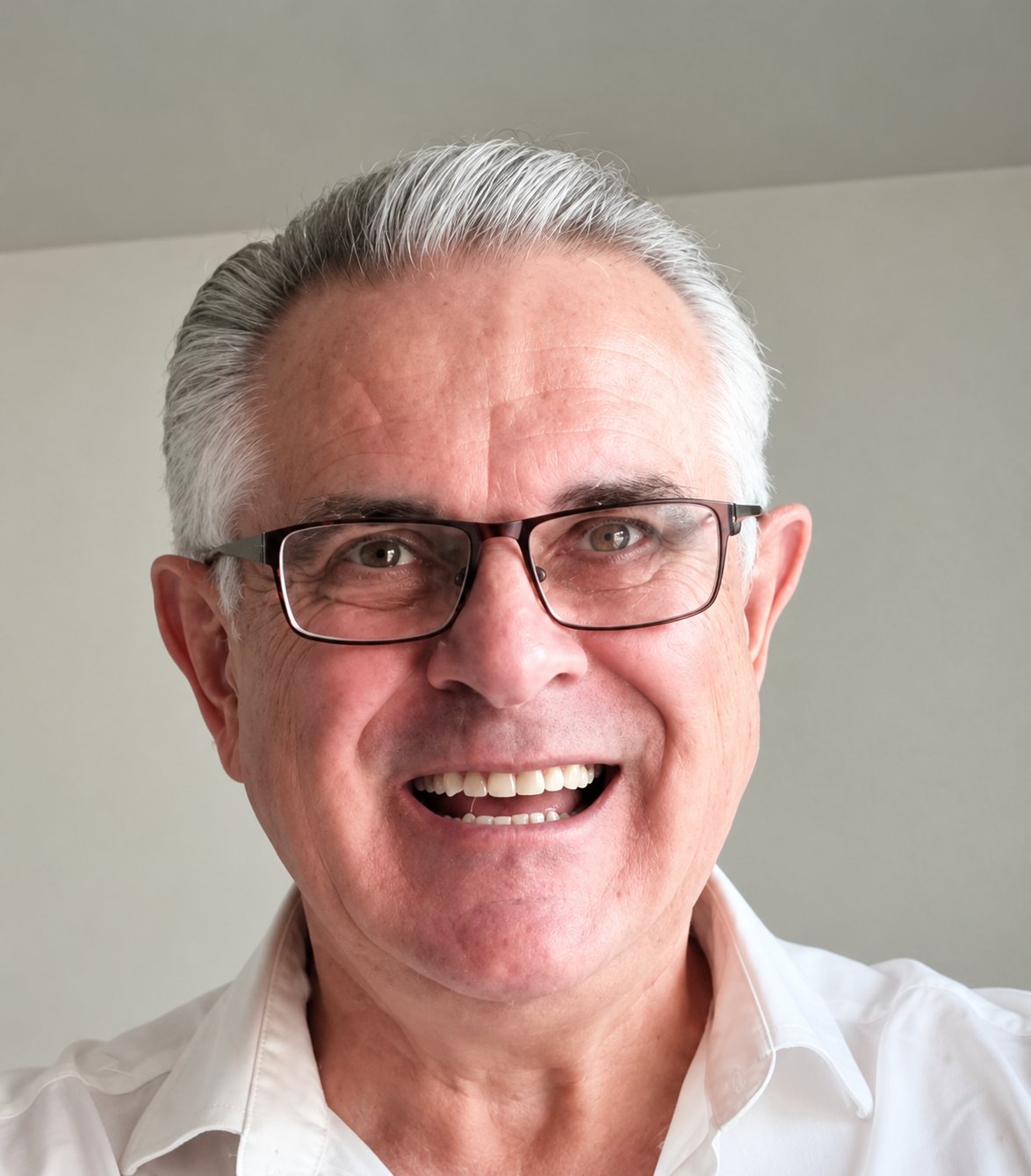 Un homme âgé aux cheveux gris et portant des lunettes sourit largement devant un mur clair, vêtu d'une chemise blanche.